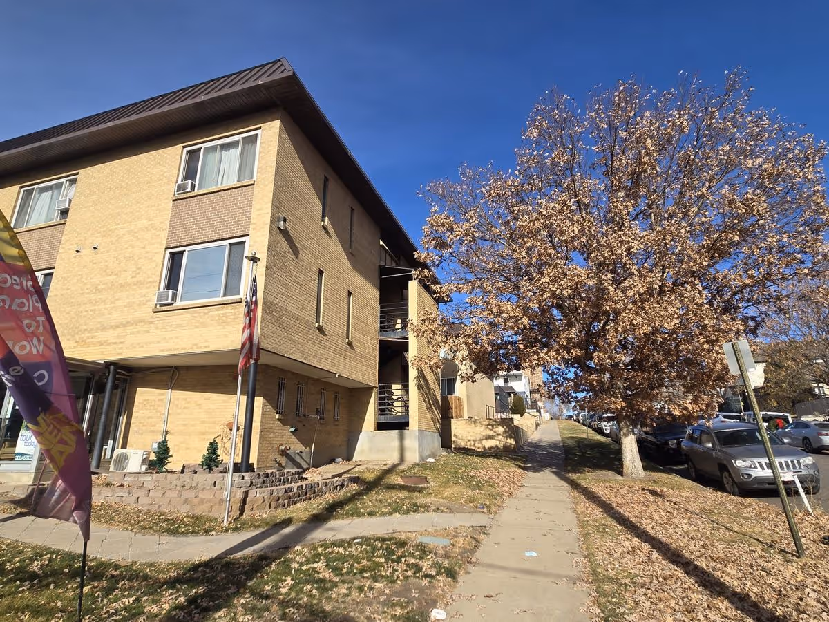 Brick multi-story building with an American flag beside a sidewalk and a large tree with brown leaves under a clear blue sky.