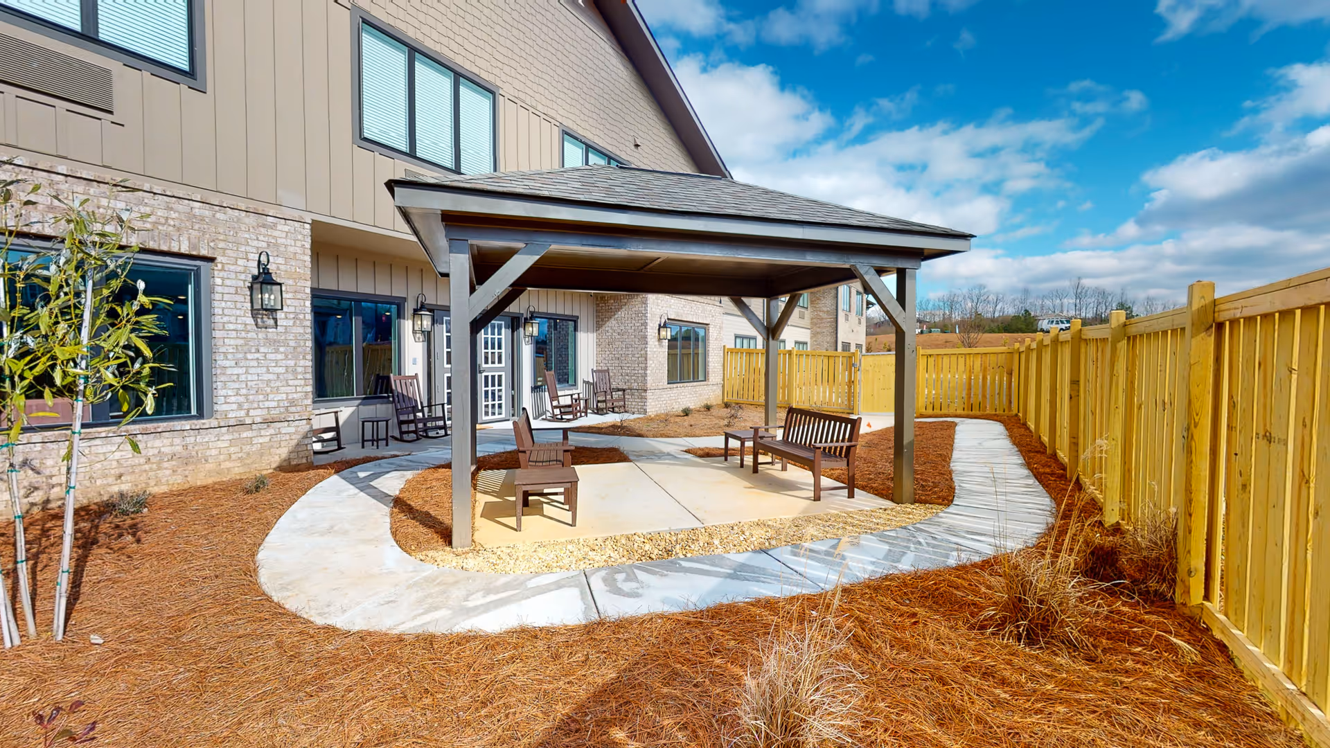 Outdoor patio area at The Lodge at Stephens Lake senior living facility featuring a covered seating area with wooden chairs and benches, a curved concrete walkway, a wooden fence, and a partly cloudy blue sky.