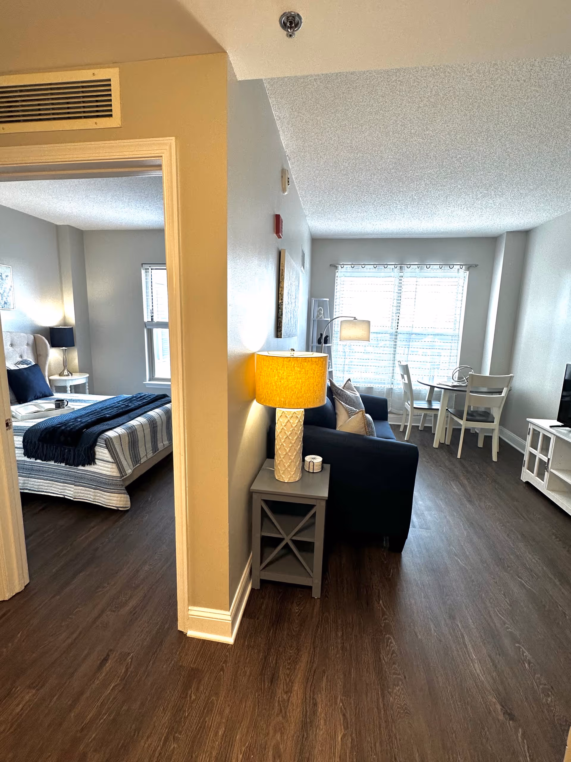 View of a senior living apartment showing a bedroom on the left with a bed, nightstand, and lamp, and a living room area on the right with a sofa, side table with a lamp, dining table with chairs, and a TV stand. The space has wood flooring and large windows with blinds letting in natural light.