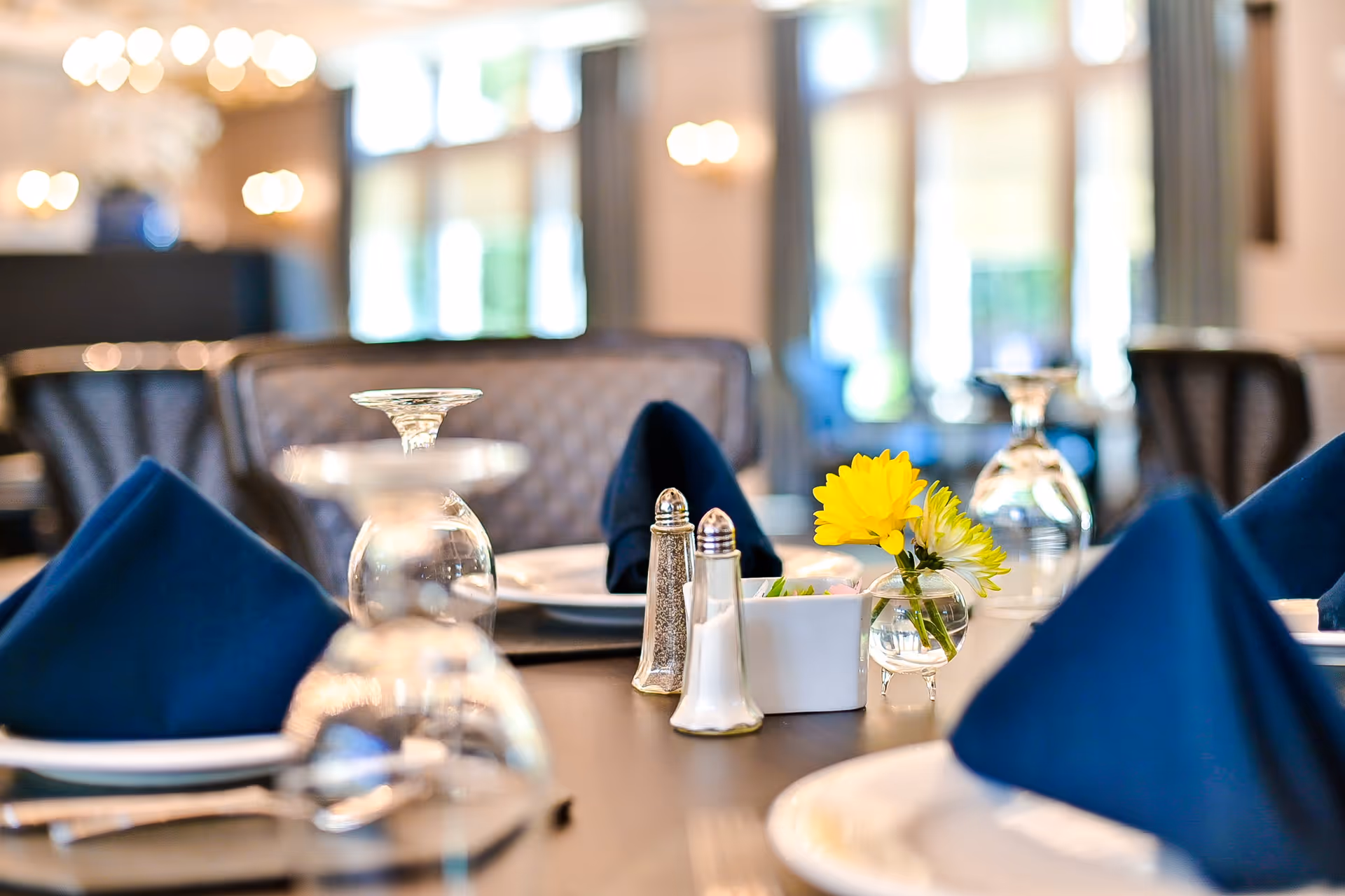 Close-up view of a dining table set with inverted wine glasses, blue folded napkins, white plates, salt and pepper shakers, a small white container, and a small vase with yellow and white flowers in a softly lit dining room.