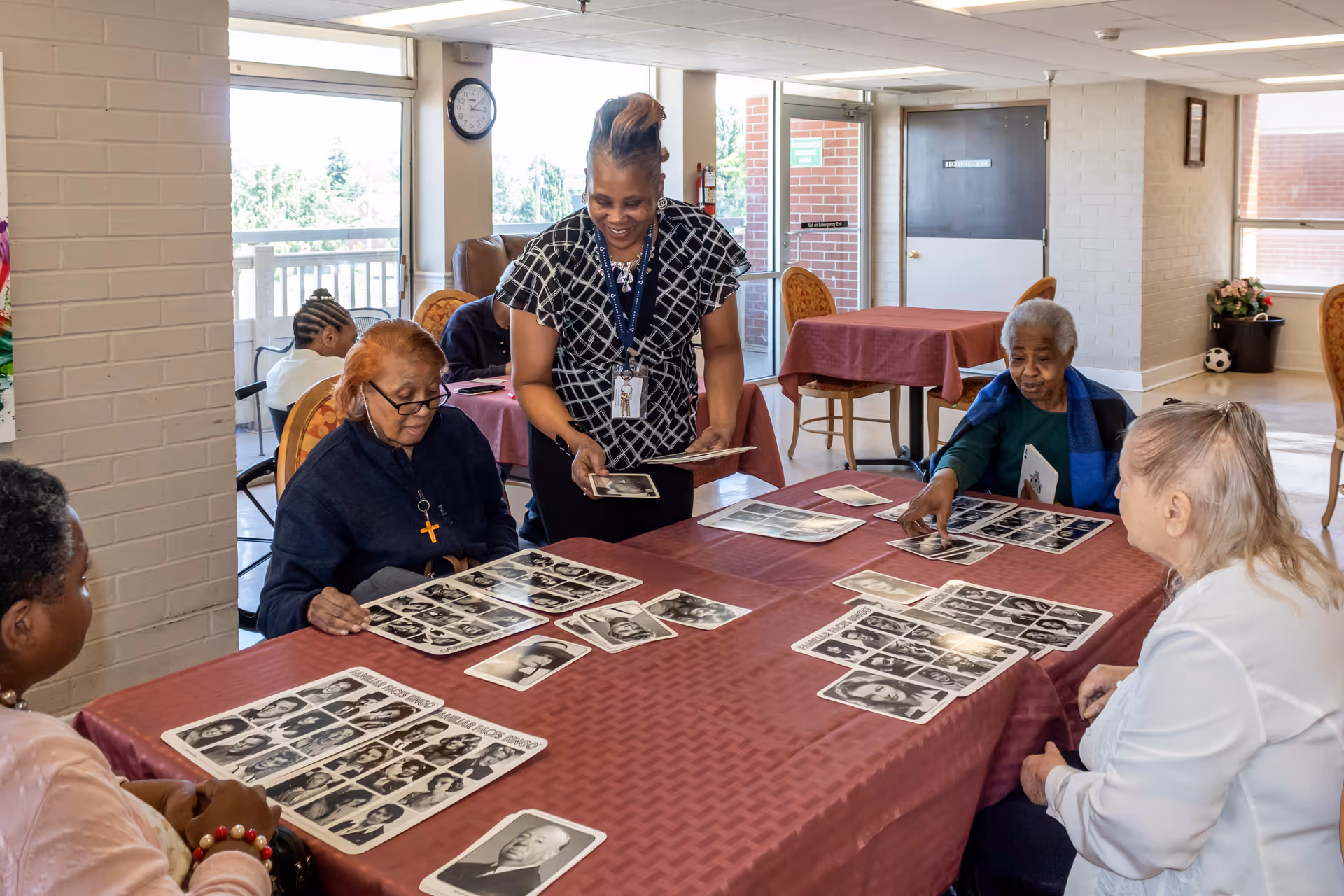 A group of elderly women sitting around a table covered with a red tablecloth, looking at black and white photographs. A staff member is standing and engaging with them, holding some photos. The room has large windows letting in natural light and a clock on the wall.
