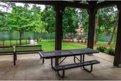 Covered outdoor picnic area with metal tables and benches overlooking a grassy yard and a pond with trees.