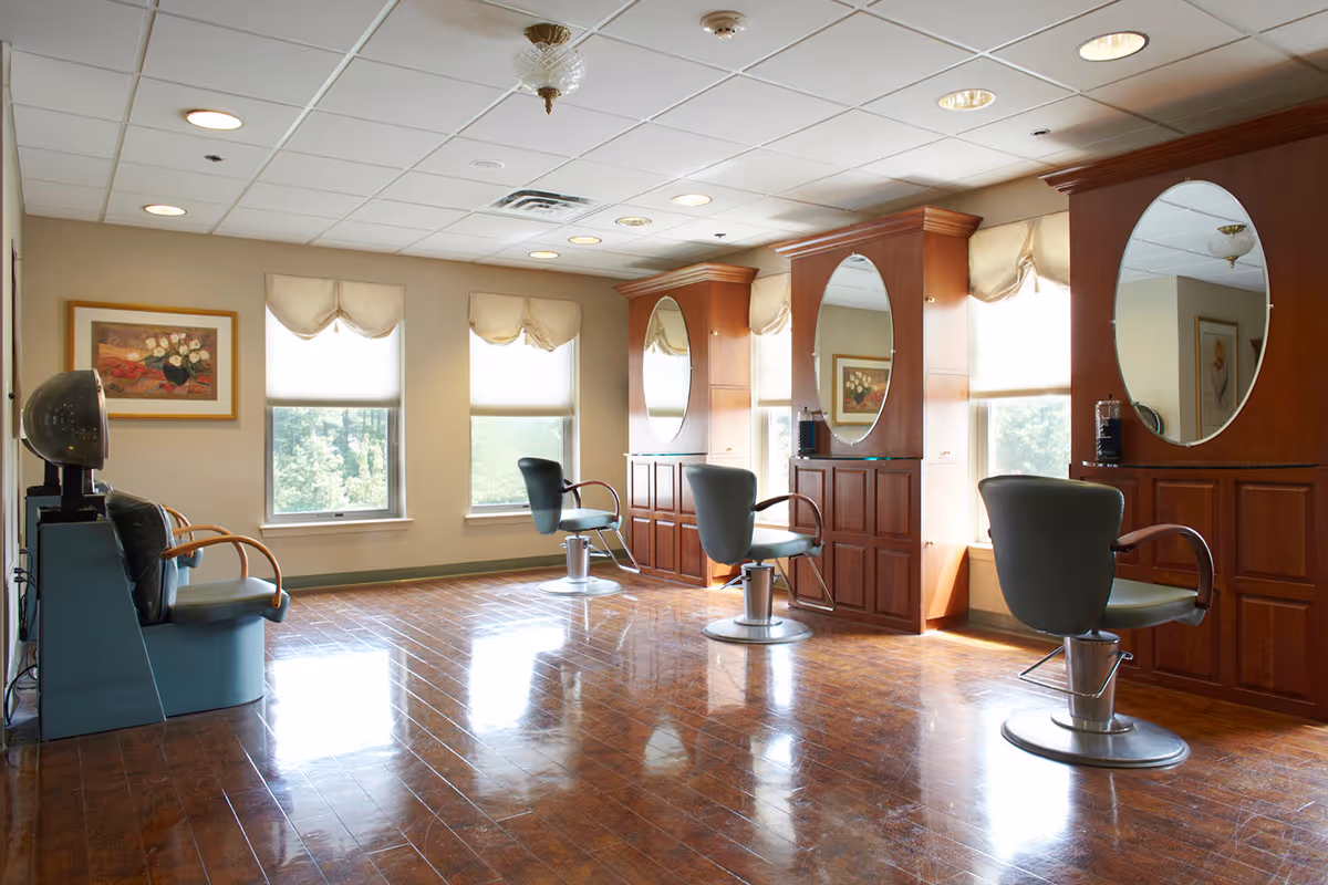 A bright and clean salon area with three styling chairs in front of large oval mirrors mounted on wooden cabinetry. The room has polished wooden floors, three windows with beige valances letting in natural light, and a hair dryer station on the left side.