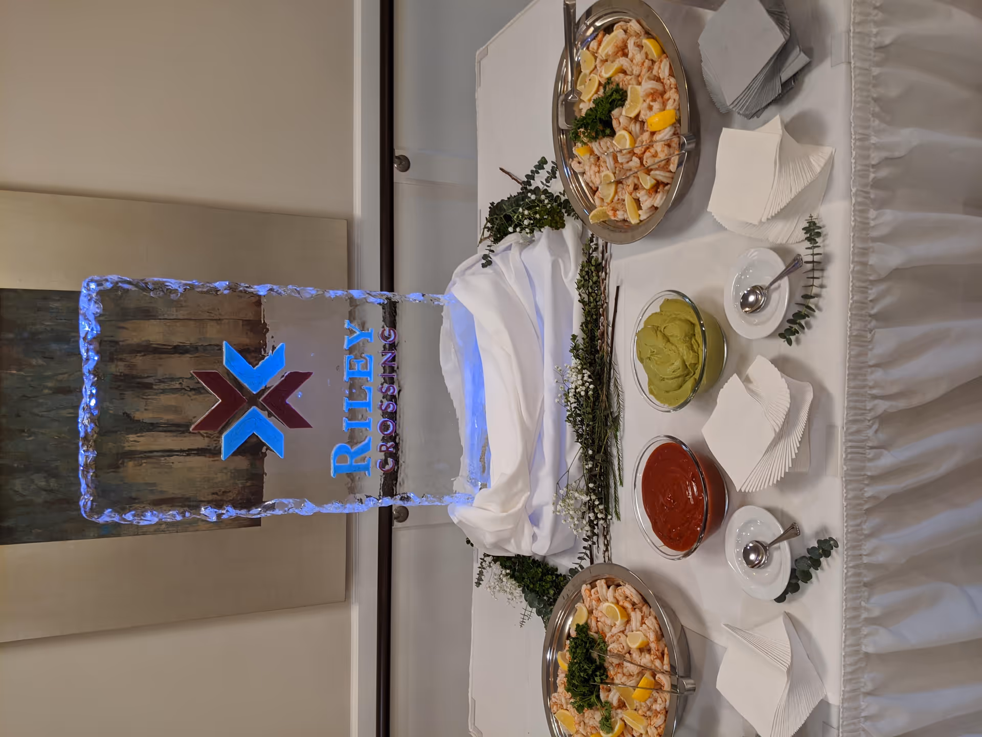 A buffet table with two platters of shrimp garnished with lemon wedges and parsley, bowls of guacamole and cocktail sauce, stacks of napkins, and an ice sculpture with the Riley Crossing Senior Living logo.