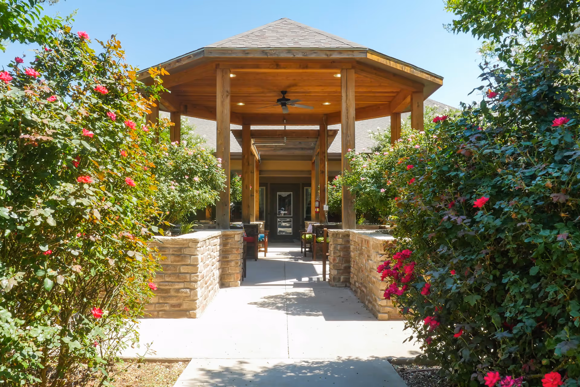 A covered outdoor walkway with a wooden roof supported by wooden pillars, flanked by stone half-walls and vibrant flowering bushes on both sides, leading to a building entrance under a clear blue sky.