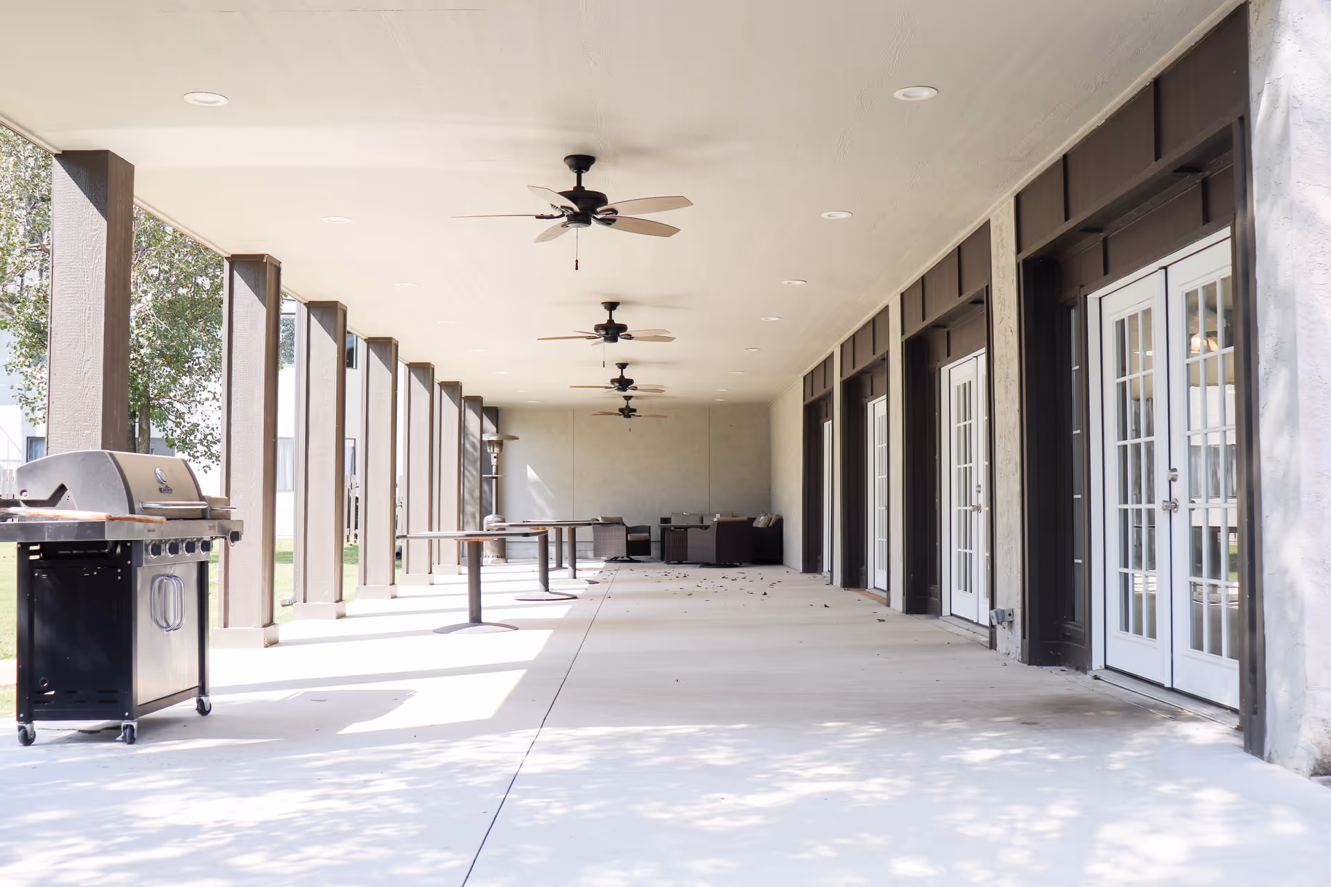 Covered outdoor patio with ceiling fans, tables, a grill, and seating along a building with glass doors.