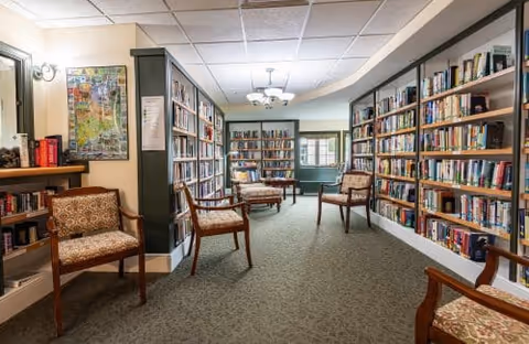Interior view of a cozy library room with bookshelves lining both walls filled with books. Several wooden chairs with patterned cushions are arranged along the room. A map is visible on the left wall, and a chandelier-style light fixture hangs from the ceiling.