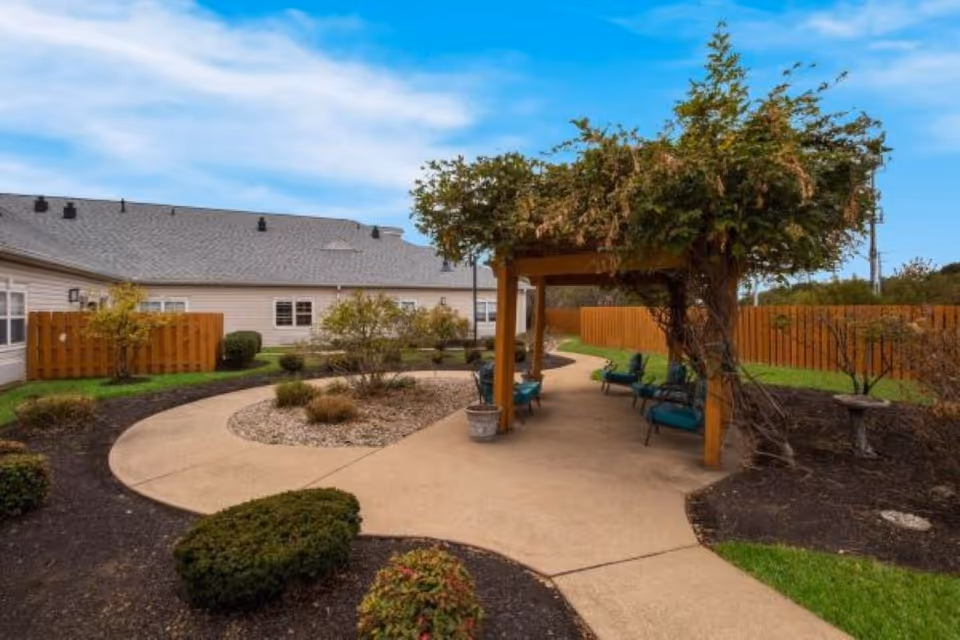 Outdoor garden area at Vitality Living Stony Brook featuring a curved concrete pathway, a wooden pergola with climbing plants, several green cushioned chairs, landscaped bushes, and a wooden fence surrounding the space under a partly cloudy blue sky.