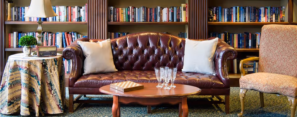 A cozy living room area featuring a dark brown leather tufted sofa with two white pillows, a wooden coffee table with a chessboard and several glass goblets, a side table covered with a patterned cloth holding a small potted plant, a vintage radio, and a lamp. Behind the sofa is a large bookshelf filled with books.