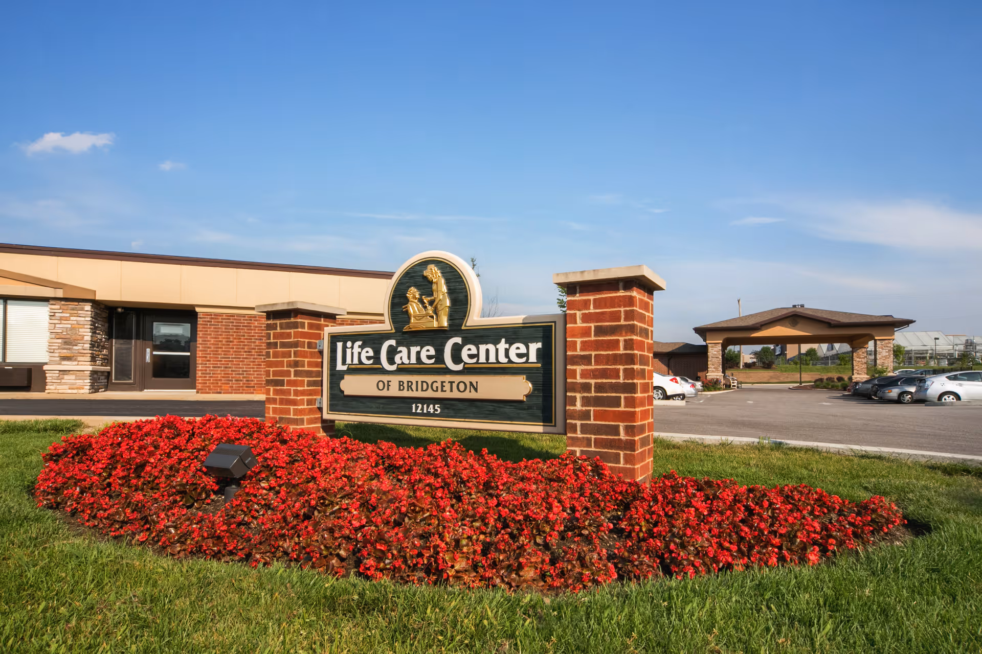 Exterior view of Life Care Center of Bridgeton with a brick sign surrounded by red flowers and green grass. The building and parking lot with several cars are visible in the background under a clear blue sky.