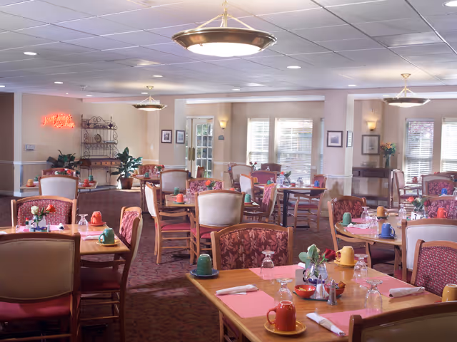 A bright and spacious dining room with multiple wooden tables and chairs arranged neatly. Each table is set with colorful mugs, glasses, napkins, and small flower arrangements. The room has carpeted floors, soft lighting from ceiling fixtures, and large windows allowing natural light to enter. There are plants and framed pictures on the walls, creating a welcoming atmosphere.