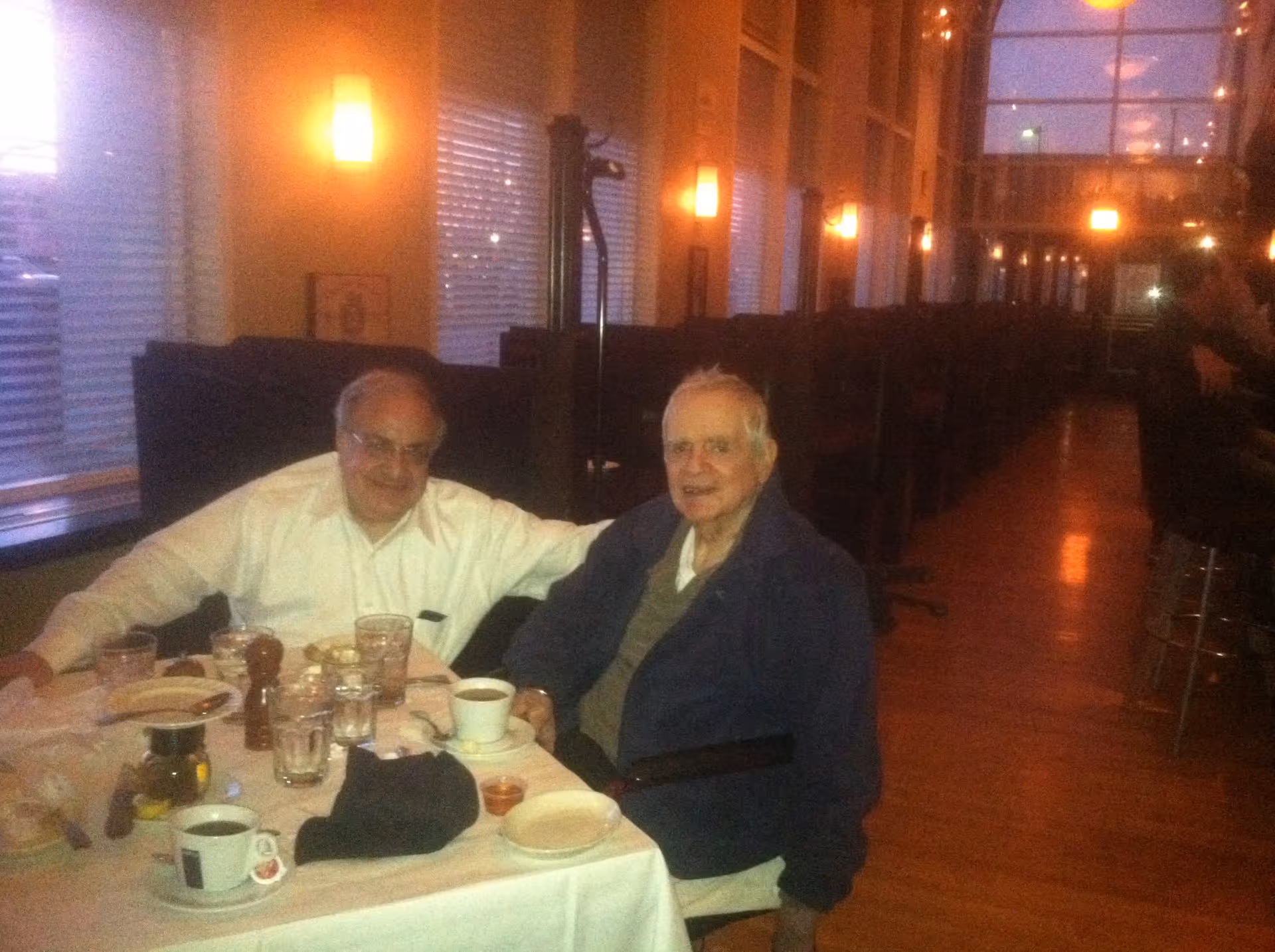 Two elderly men sitting at a table in a warmly lit restaurant or dining area with large windows and booth seating along the wall. The table has cups, glasses, plates, and condiments on it.