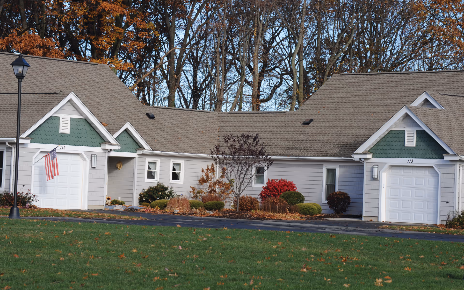 Single-story connected cottage-style residences with attached garages, landscaped front beds, and autumn trees in the background.
