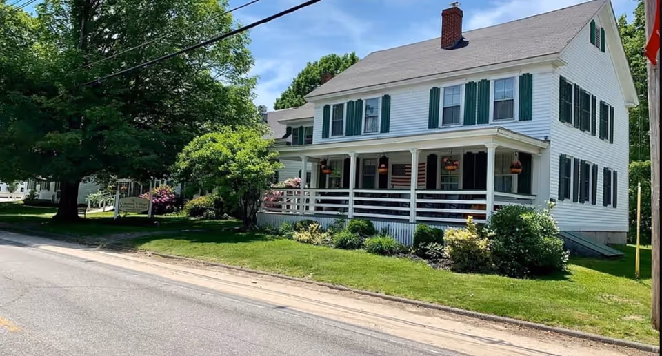 A large white two-story house with green shutters and a porch with hanging flower baskets, surrounded by green grass, bushes, and trees on a sunny day.