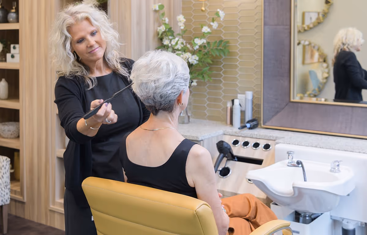 A stylist trims an older woman's short gray hair in a bright salon chair beside a wash sink and mirror.