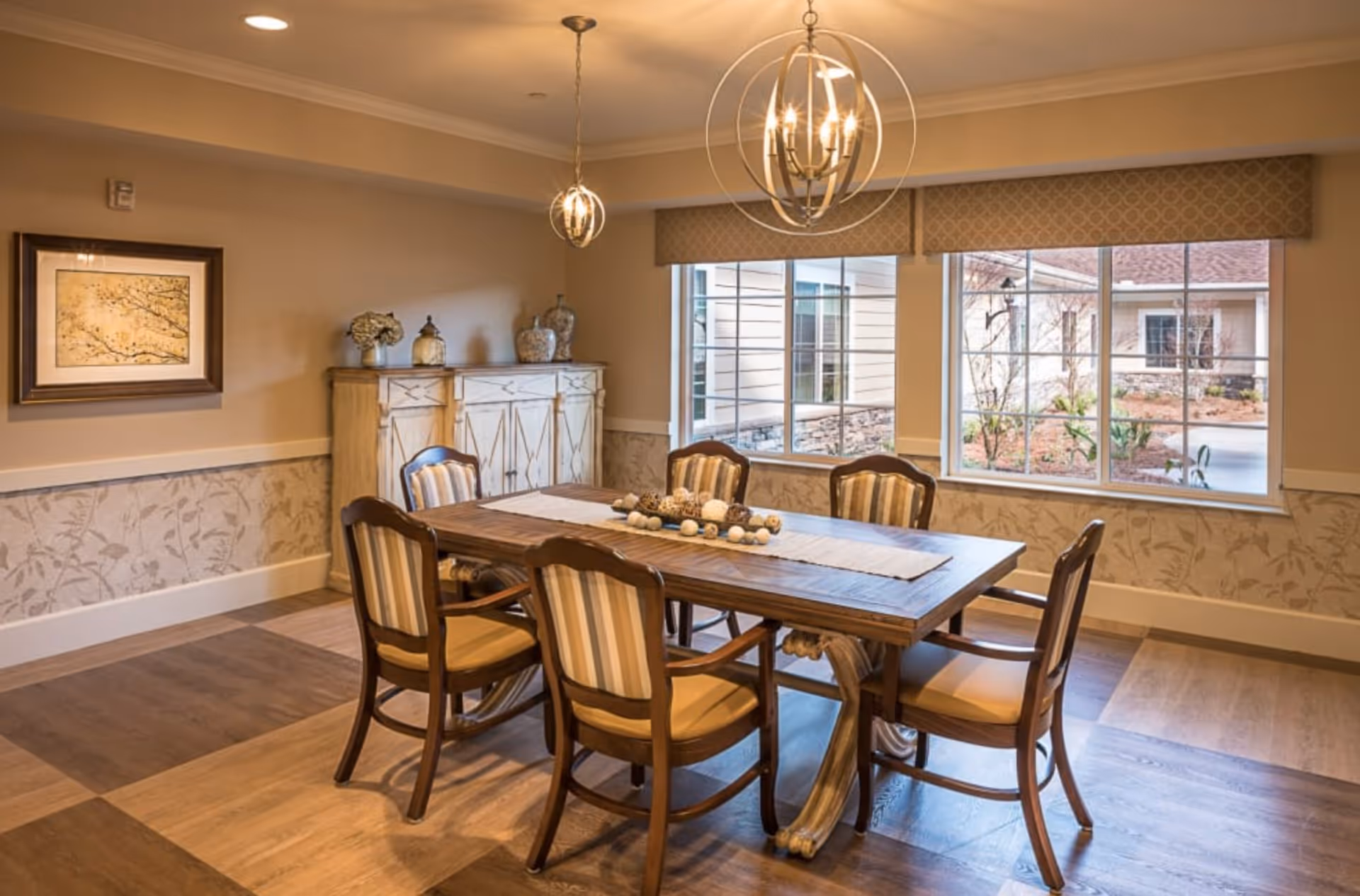 A well-lit dining room with a wooden table surrounded by six cushioned chairs with striped upholstery. The table has a decorative centerpiece with assorted natural elements. Behind the table is a wooden sideboard with decorative vases and a framed artwork on the wall. Large windows with patterned valances allow natural light to enter, showing an outdoor courtyard area.