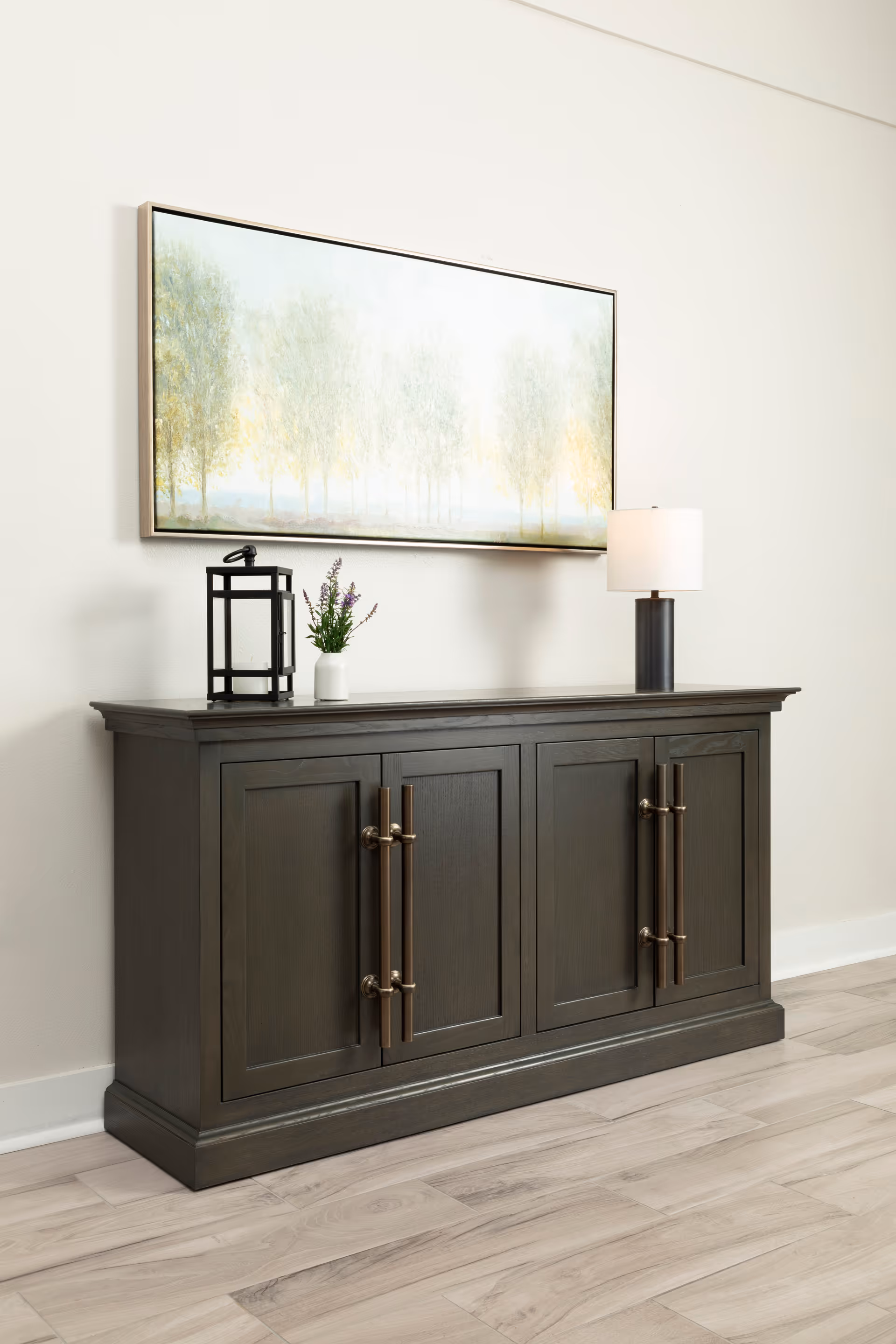 A dark wooden sideboard with four cabinet doors and bronze handles sits against a white wall. On top of the sideboard are a black lantern, a small white vase with purple flowers, and a modern table lamp with a white shade. Above the sideboard hangs a large rectangular painting depicting a soft, misty forest scene with trees in light green and yellow tones. The floor is light wood.