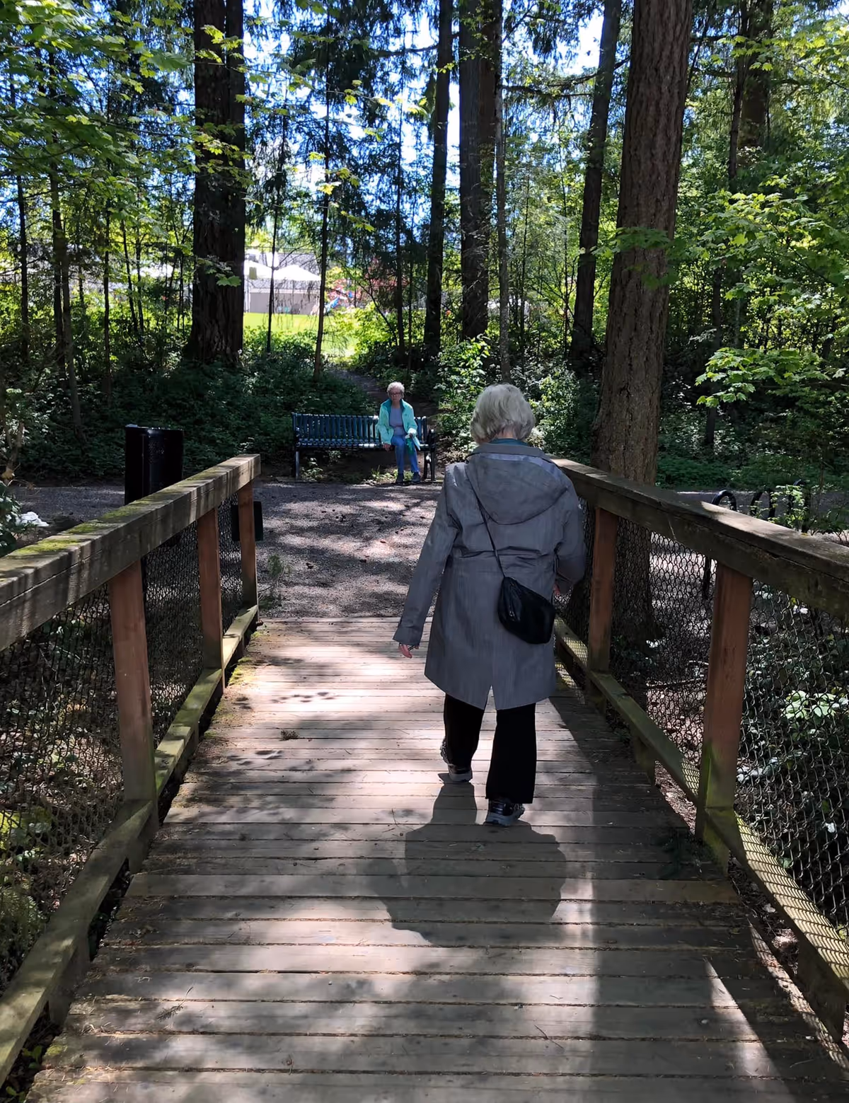 An elderly woman wearing a gray coat and carrying a black purse walks across a wooden bridge in a wooded outdoor area. Another elderly woman is sitting on a bench in the background surrounded by trees and greenery.