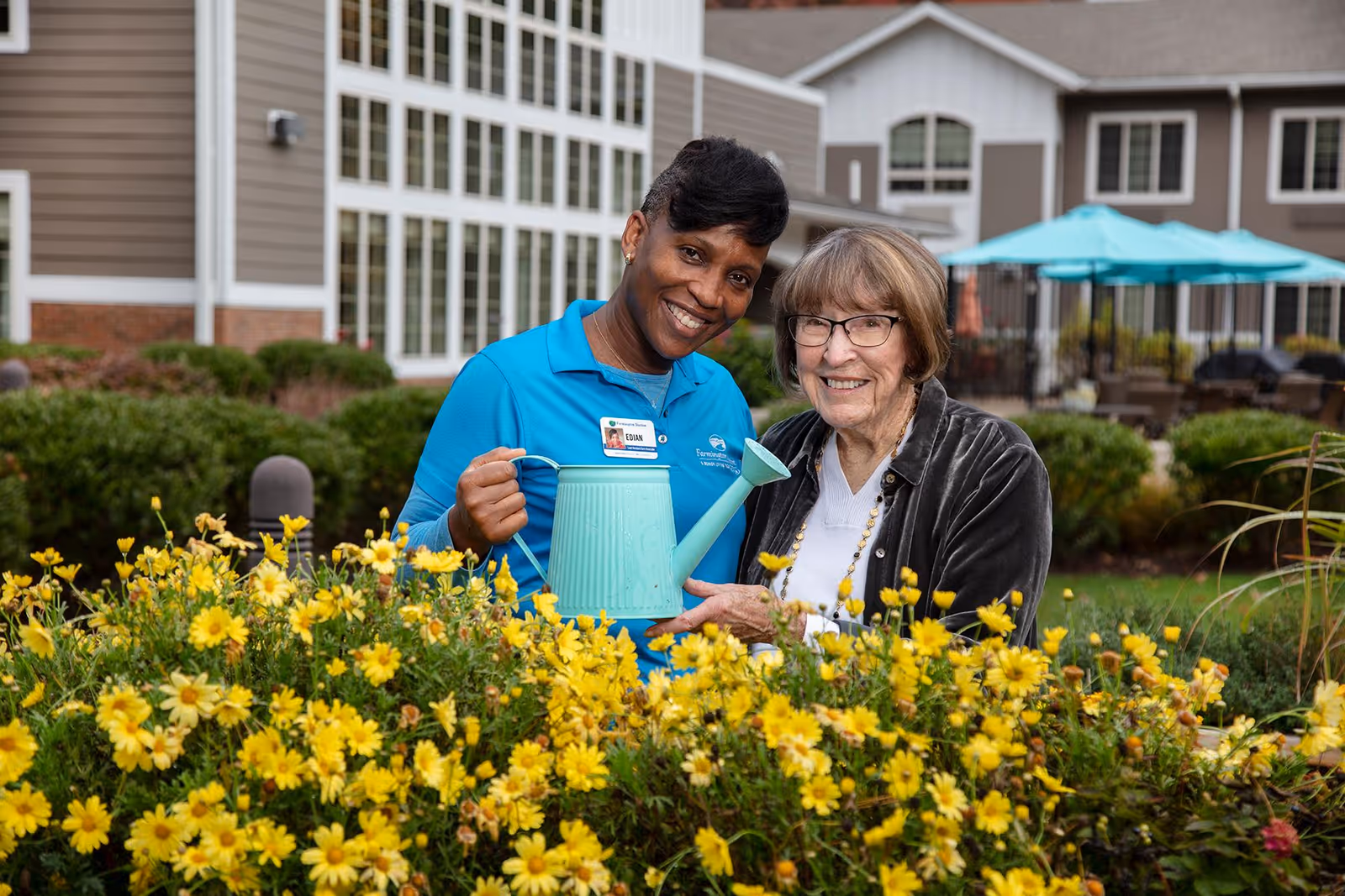 A smiling caregiver and an elderly woman stand together outdoors in a garden with yellow flowers. The caregiver is holding a light blue watering can, and both are looking at the camera. Behind them is a senior living facility building with large windows and outdoor seating with blue umbrellas.
