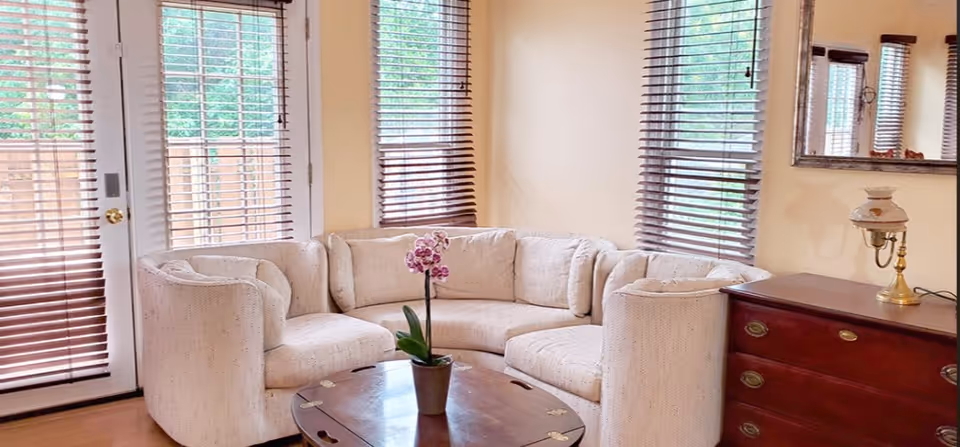 Sunlit living room featuring a curved beige sectional sofa, a round wooden coffee table with a potted orchid, wooden blinds, and a dresser with a lamp.
