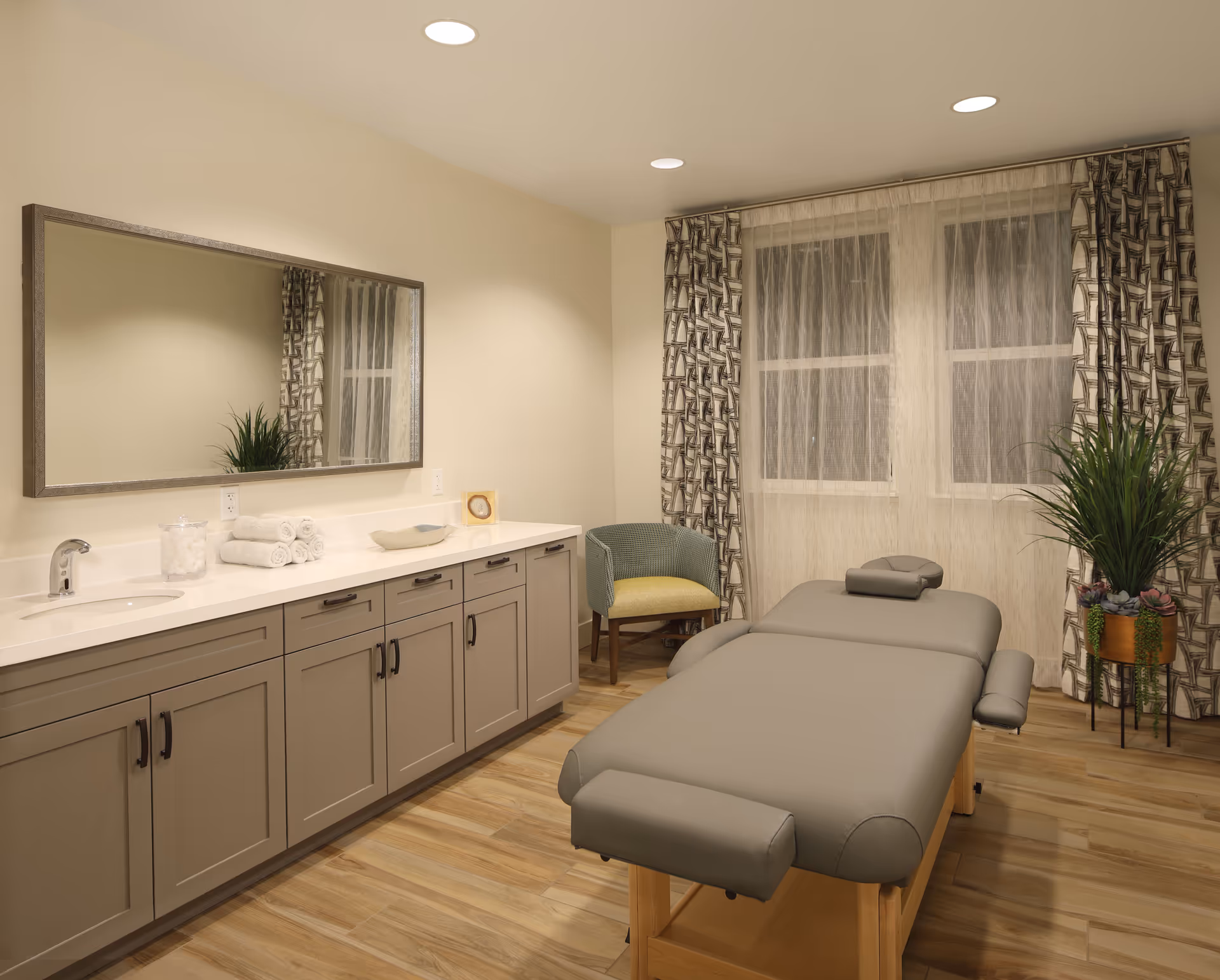 A serene room with a massage table in the center, a large mirror above a long cabinet with a sink on the left, a green cushioned chair near the window with patterned curtains, and a potted plant on the right. The room has wooden flooring and recessed ceiling lights.