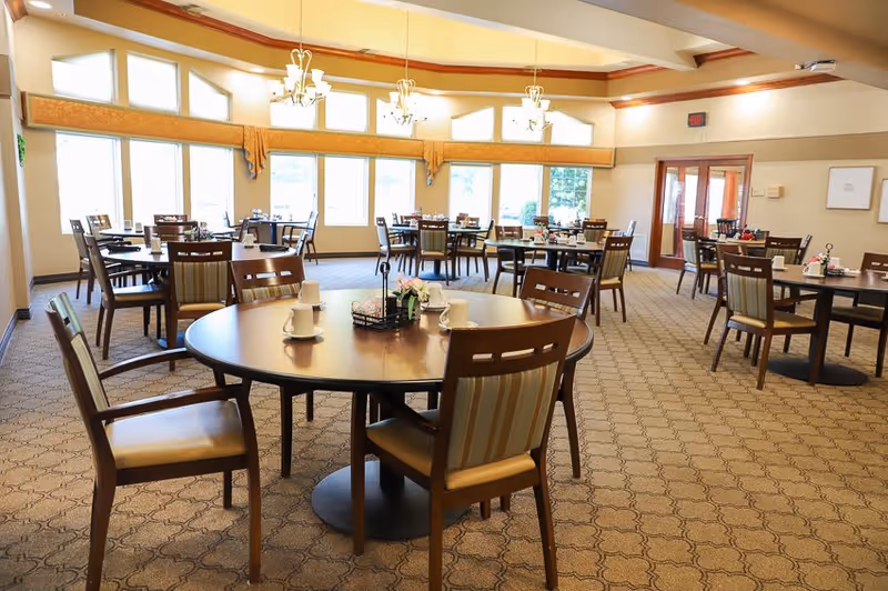 A spacious dining room with multiple round tables and wooden chairs with cushioned seats. Each table has cups, napkins, and small flower arrangements. Large windows with valances allow natural light to fill the room, and chandeliers hang from the ceiling. The room has a patterned carpet and light-colored walls.