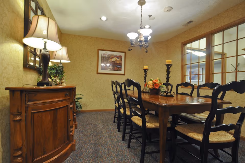 A dining room with a long wooden table surrounded by eight black chairs with woven seats. On the table, there are two tall candle holders with yellow candles and a floral centerpiece. To the left, there is a wooden sideboard with a lamp and a mirror above it. The walls are covered with patterned wallpaper, and a framed picture hangs on the far wall. A chandelier with five lights hangs from the ceiling, and a large window with wooden frames looks into another room.