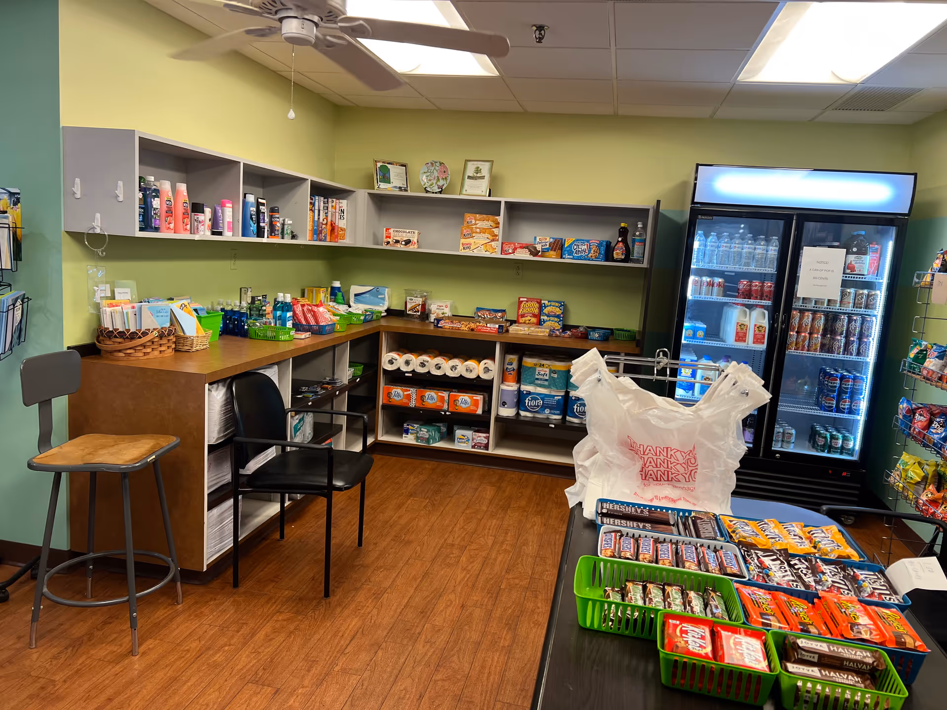 Interior of a small convenience store or snack shop with shelves stocked with snacks, toiletries, paper products, and a refrigerated display case with drinks. There are two chairs near the counter and a ceiling fan above.