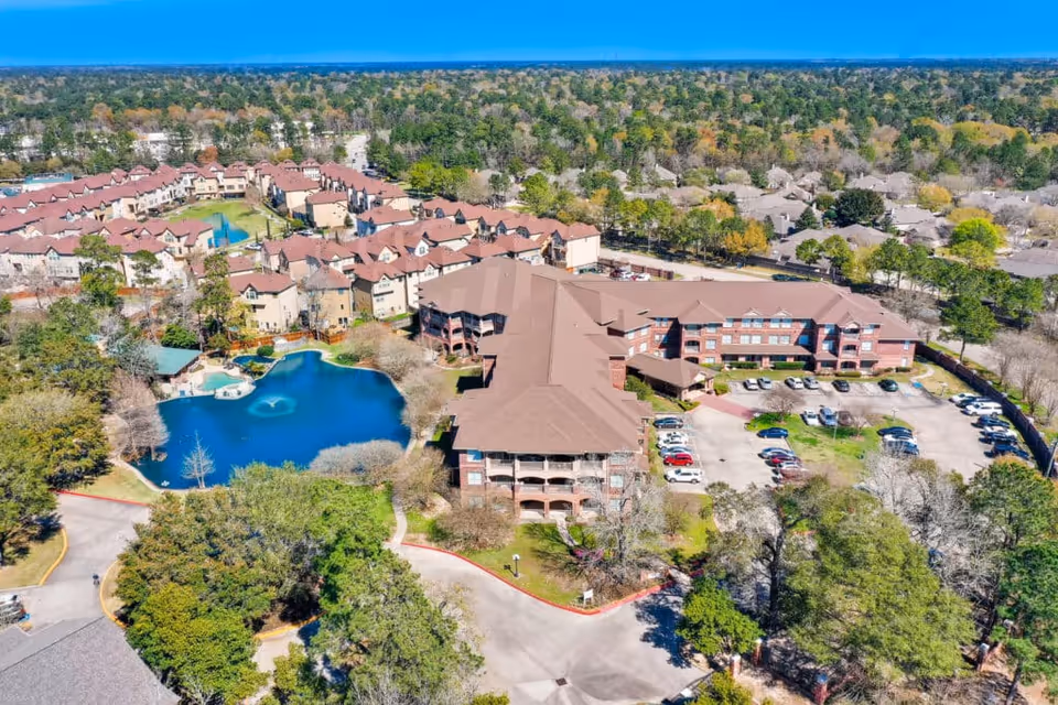Aerial view of The Terraces at Kingwood Town Center senior living facility showing multiple buildings with brown roofs, a large pond with a fountain, surrounding trees, parking lots with cars, and a residential neighborhood in the background under a clear blue sky.