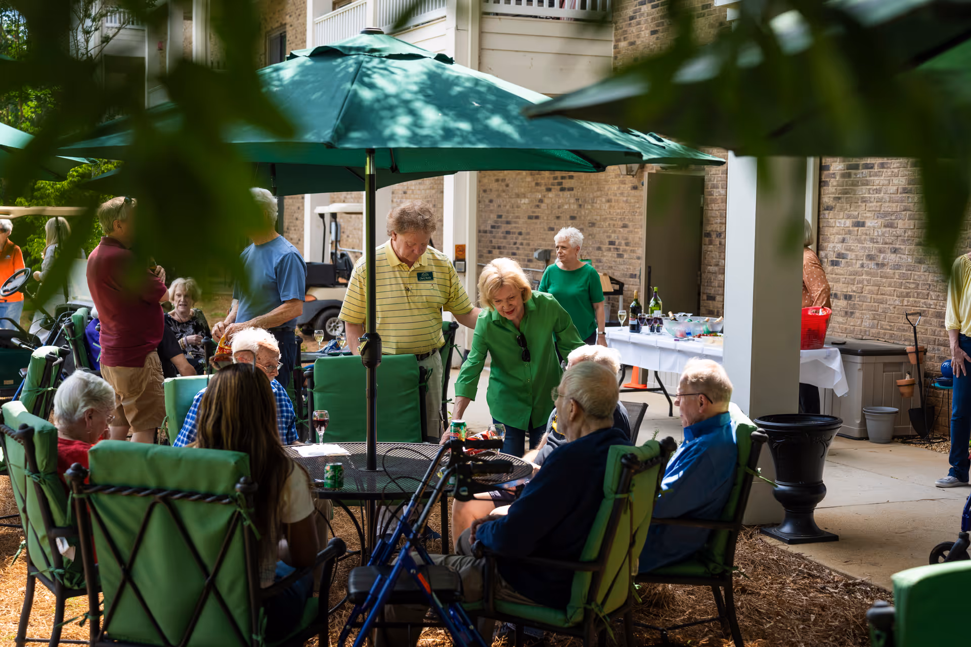 A group of elderly people socializing outdoors at a senior living facility. They are seated around a table with green cushioned chairs under a large green patio umbrella. Some people are standing and chatting near a table with drinks and snacks in the background. The setting is a courtyard area with brick walls and greenery.
