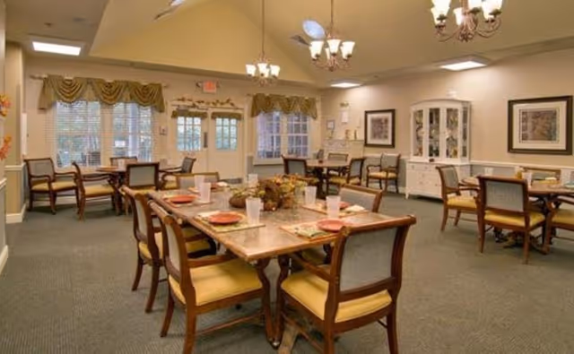 A spacious dining room with multiple wooden tables and chairs arranged neatly. Each table is set with plates, napkins, and cups. The room has large windows with decorative curtains, chandeliers hanging from the ceiling, and framed artwork on the walls. There is a white cabinet against one wall and a door leading outside.
