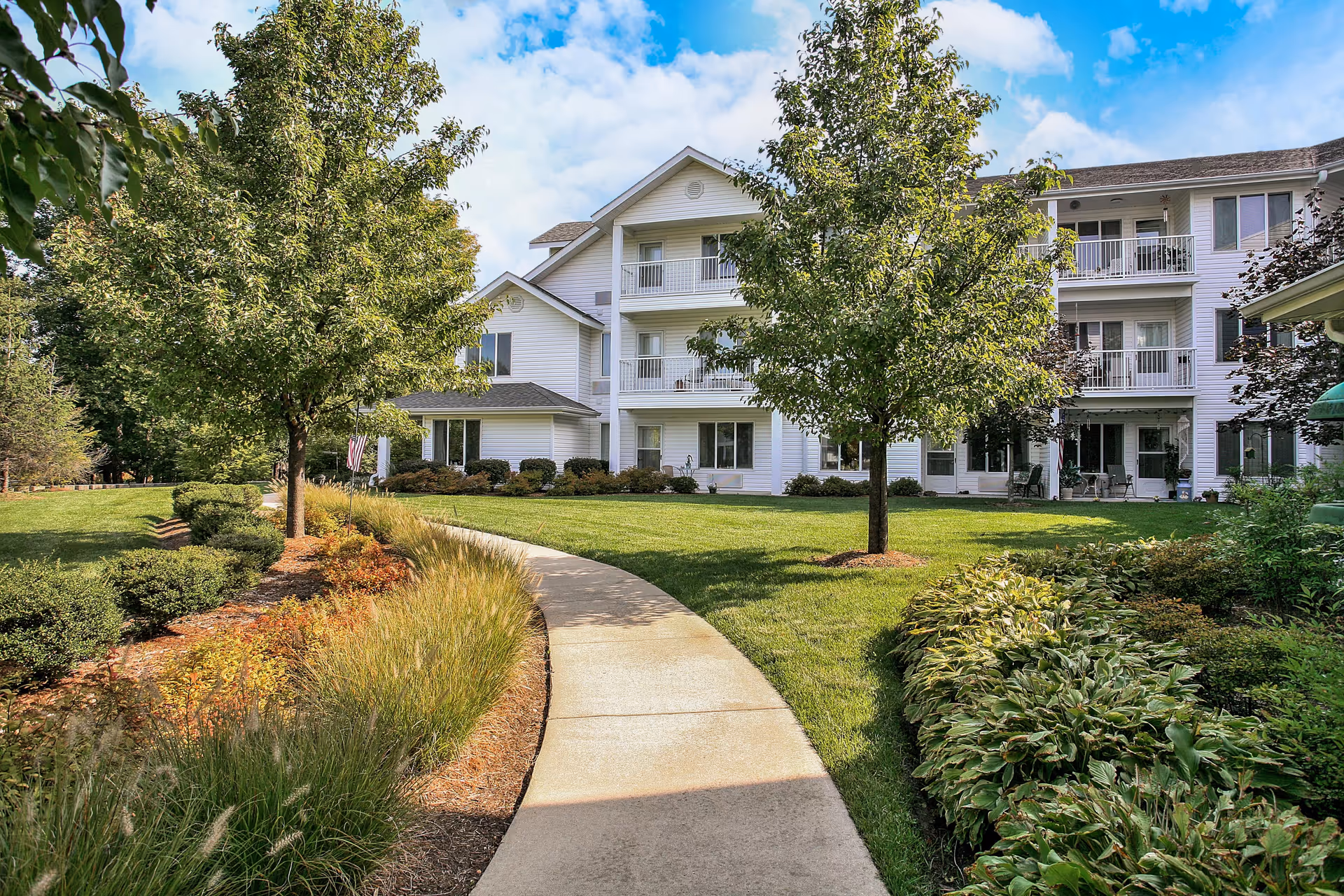 A paved walkway curves through a landscaped garden area with green grass, bushes, and trees leading to a white multi-story residential building with balconies under a partly cloudy blue sky.