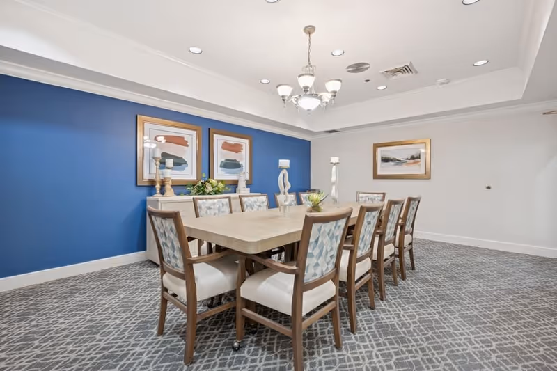 A well-lit dining room with a rectangular table surrounded by ten cushioned chairs with patterned backs. The room features a blue accent wall with two abstract paintings and decorative candle holders on a sideboard. A chandelier hangs from the ceiling, and the floor is carpeted with a geometric pattern.