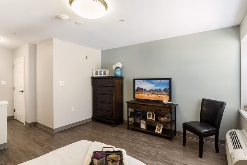 A cozy living area in a senior living facility featuring a wooden dresser with a vase of white flowers and framed photos on top, a TV on a wooden stand displaying a desert mountain scene, a black leather chair, and a window air conditioning unit. The room has light-colored walls and wood flooring.