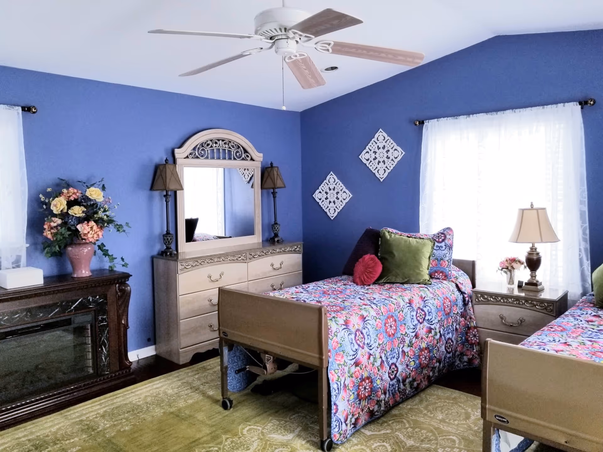 A bright bedroom with two single beds covered in colorful floral bedspreads. The walls are painted blue, and there is a ceiling fan above. Between the beds is a nightstand with a lamp and a small flower arrangement. A dresser with a large mirror and two lamps is against one wall, and a decorative fireplace with a vase of flowers is on the left side. Two white decorative wall hangings are on the wall above one bed, and sheer white curtains cover the windows.