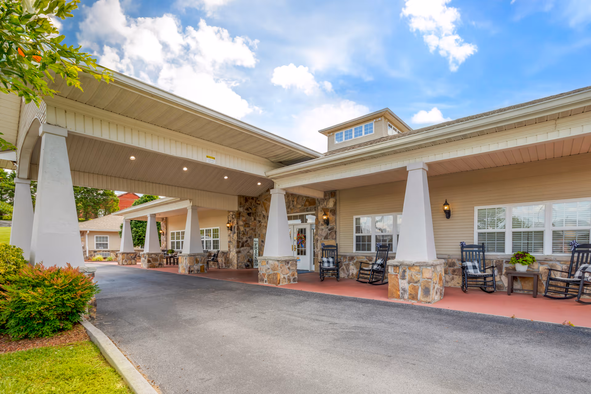 Front exterior view of a senior living facility with a covered driveway supported by large white pillars with stone bases. There are rocking chairs and small tables on the porch area, with a clear blue sky and some clouds above.