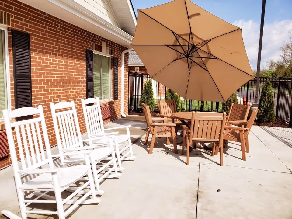 Outdoor patio area with four white rocking chairs lined up against a brick wall and a round table with six brown chairs under a large beige umbrella. There is a black metal fence and some greenery in the background under a partly cloudy sky.