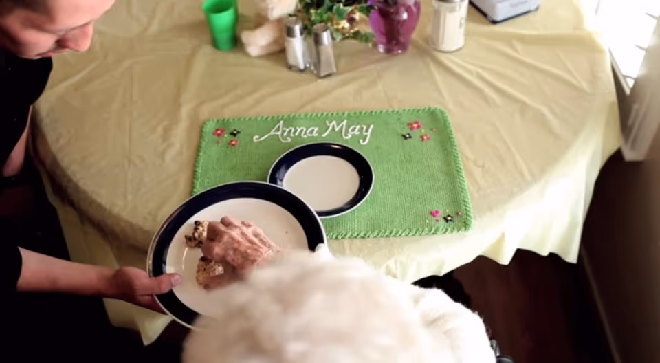 An elderly person with white hair is seated at a table with a yellow tablecloth and a green placemat embroidered with the name 'Anna May'. A caregiver or helper is holding a plate with food, assisting the elderly person who is reaching for the food.