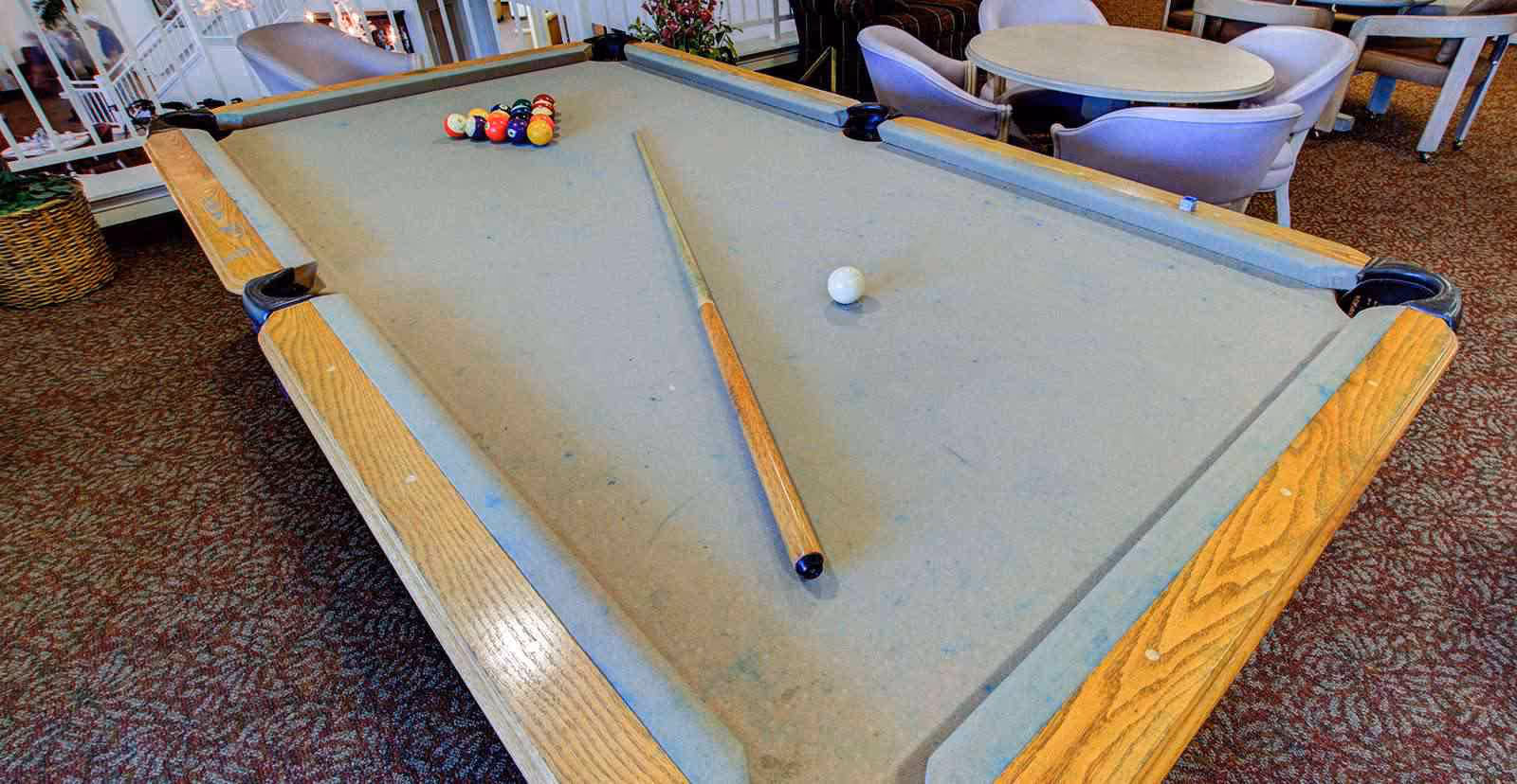 A pool table with a cue stick and billiard balls arranged in a triangle on the felt surface, located in a room with carpeted floor and round tables with chairs in the background.