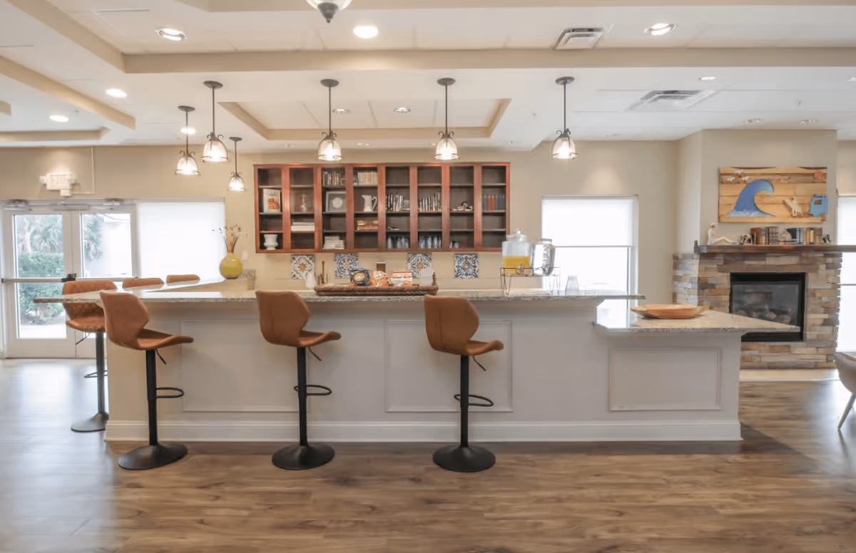 A bright and spacious kitchen area in a senior living facility featuring a long island with a granite countertop and five brown bar stools. Above the island, there are six pendant lights hanging from the ceiling. Behind the island, there are wooden cabinets with glass doors displaying books and decorative items. To the right, there is a stone fireplace with a wooden mantel decorated with books and a wooden wall art piece depicting a wave and a bird. The floor is wooden, and there are large windows allowing natural light to fill the room.