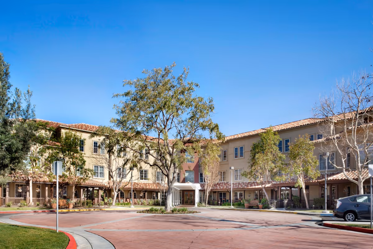 Exterior view of a three-story senior living facility building with beige walls and red-tiled roof under a clear blue sky. There are trees and shrubs in front of the building, a circular driveway, and a parked car on the right side.