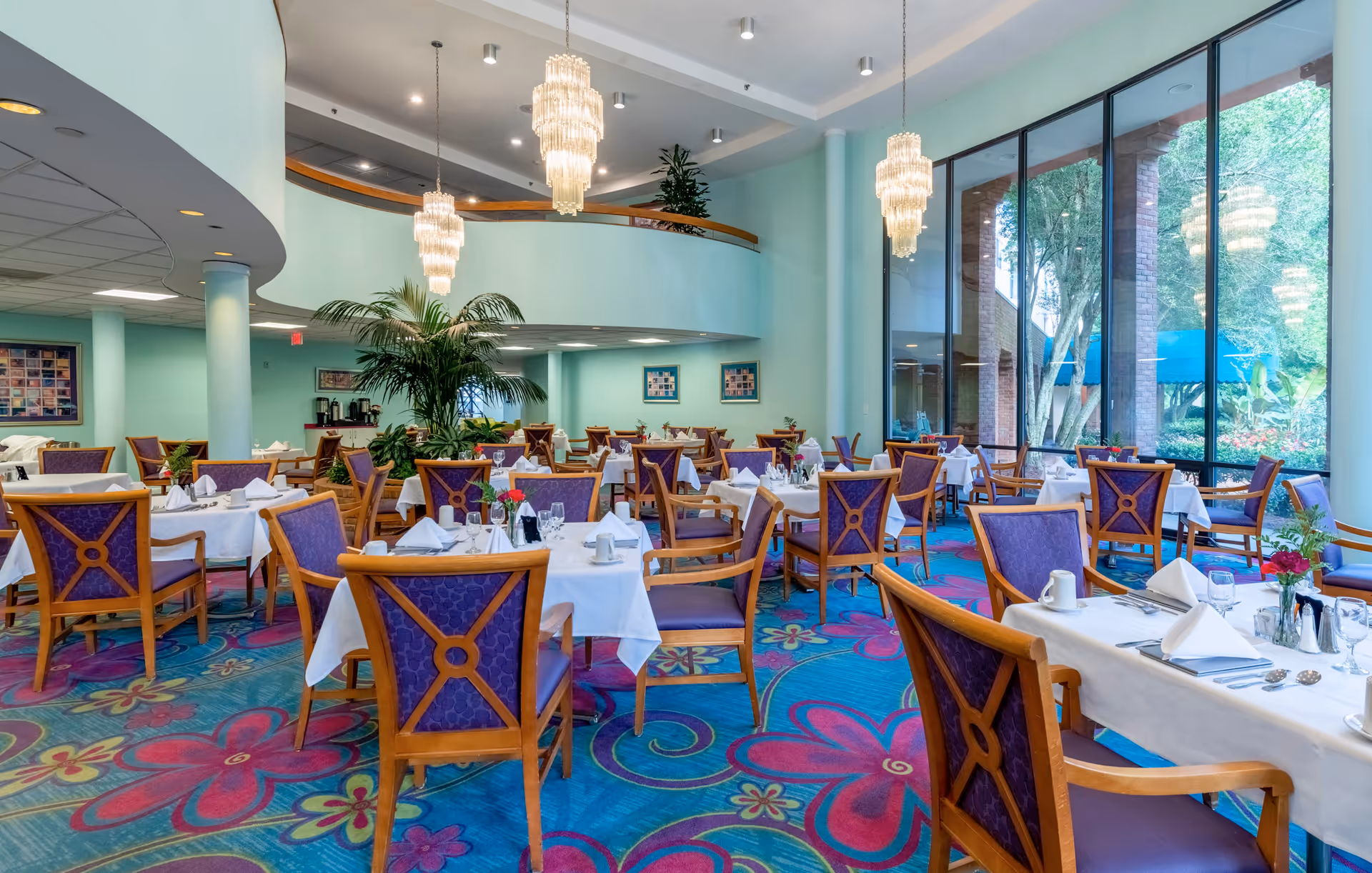 A bright and spacious dining room with multiple tables covered in white tablecloths, set with napkins, glasses, and silverware. The chairs have wooden frames with purple upholstery. The room features large floor-to-ceiling windows letting in natural light and offering a view of greenery outside. There are three elegant chandeliers hanging from the ceiling and a colorful carpet with floral patterns covering the floor.