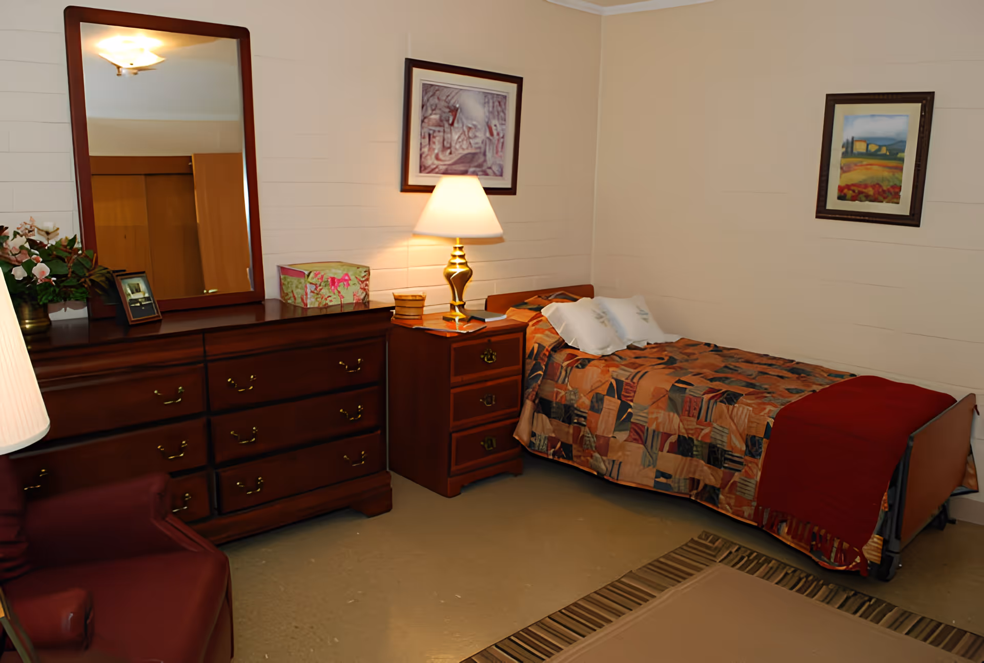 A furnished single bedroom with a patterned bed, wooden dresser and nightstand with a lamp, framed pictures on the walls, and an armchair.