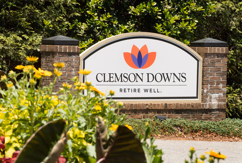 Entrance sign reading "Clemson Downs — Retire Well." mounted on a brick base with flowers in the foreground.