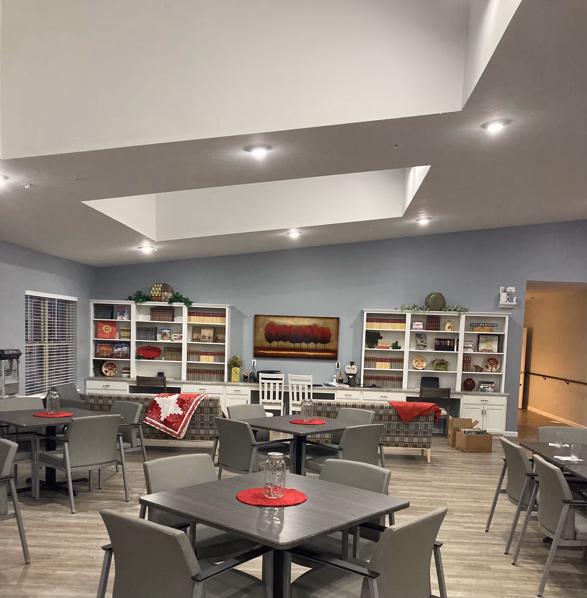 A spacious common area with multiple square tables and chairs arranged for seating. The room has light wood flooring, light blue walls, and a ceiling with recessed lighting and skylights. At the back, there are two white bookshelves filled with books and decorative items, flanking a painting of red trees. Two patterned couches with red blankets are positioned in front of the bookshelves. A hallway is visible to the right.