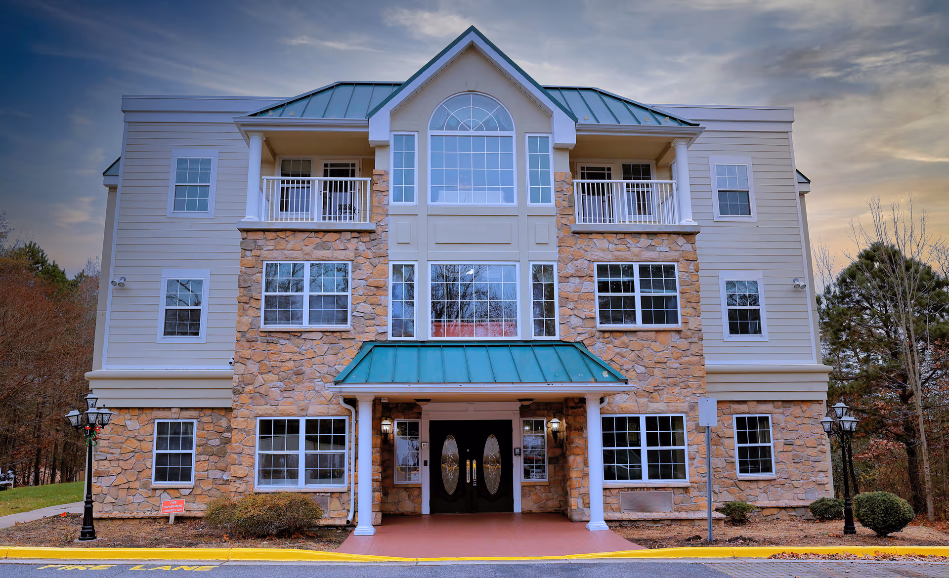 Front exterior view of a three-story assisted living facility building with stone and siding facade, green metal roof accents, multiple windows, and a covered entrance with double doors. The building is surrounded by trees and has a paved driveway with yellow curb markings.