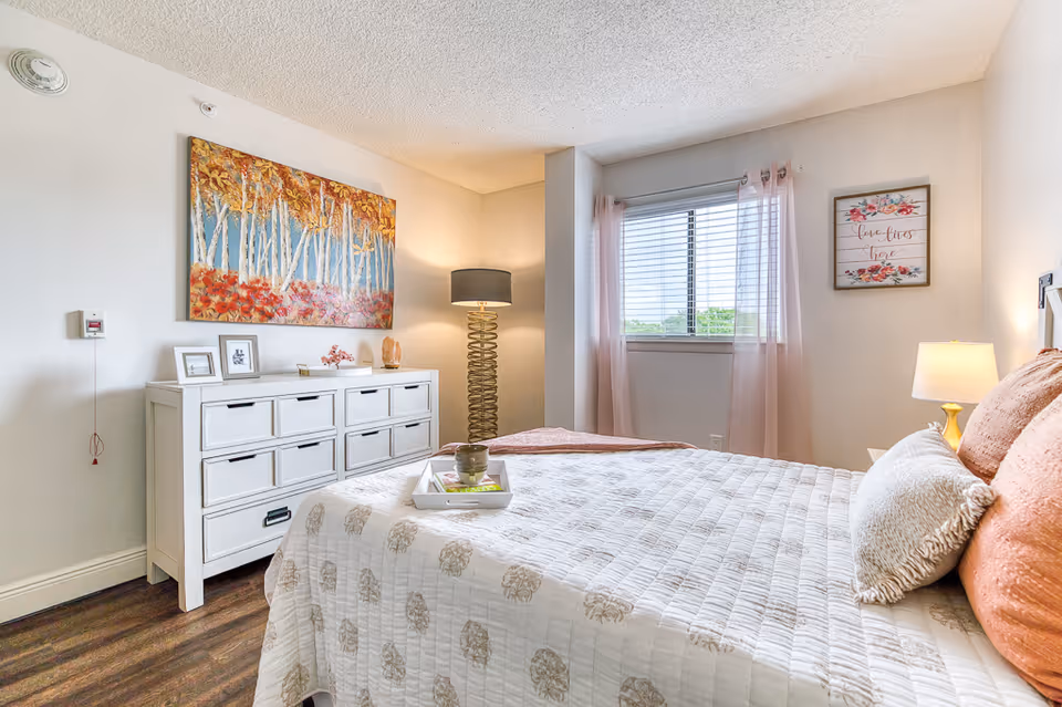 A bright and cozy bedroom with a large bed covered in a white quilt with subtle patterns. The bed has multiple pillows in shades of peach and cream. Next to the bed is a small nightstand with a lamp. Across from the bed is a white dresser with multiple drawers, decorated with framed pictures, a small plant, and a lamp with a unique spiral base. Above the dresser hangs a colorful painting of birch trees with autumn leaves. A window with sheer pink curtains lets in natural light, and a framed floral print with the words 'love lives here' hangs on the wall beside the window. The room has wooden flooring and light-colored walls.