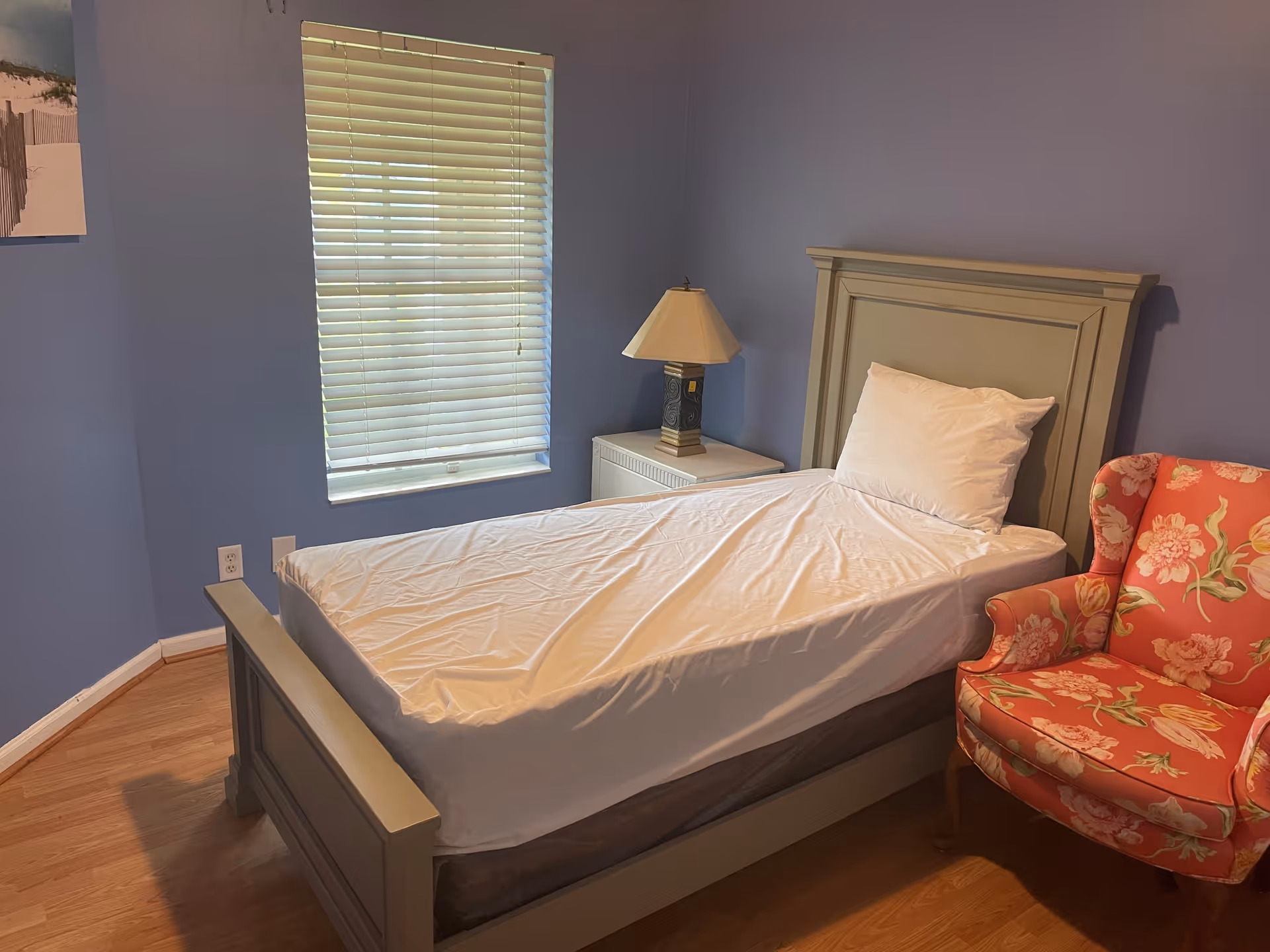 Single bed with white sheets and pillow in a blue-walled bedroom beside a nightstand with a lamp and a floral armchair.