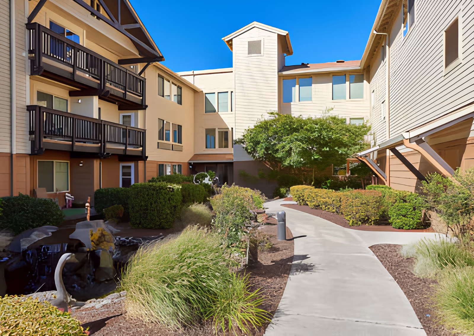 Outdoor courtyard area of Elliott Residence featuring a paved walkway surrounded by green shrubs, bushes, and a small water feature with rocks. The multi-story building with beige and brown siding has balconies overlooking the courtyard under a clear blue sky.