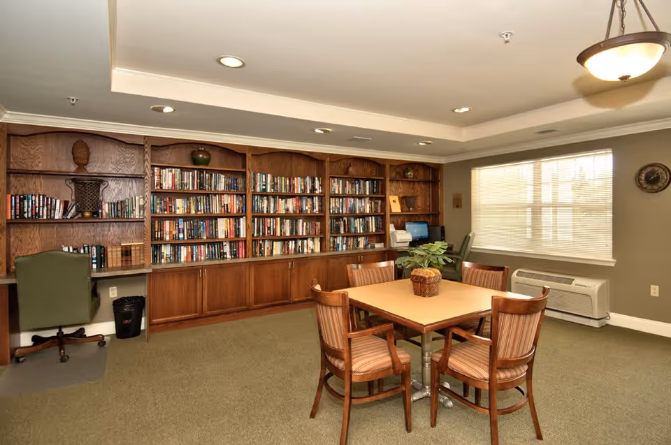 A cozy room with a wooden bookshelf filled with books along one wall, a square table with four wooden chairs in the center, a green office chair at a built-in desk with a computer, and a window with blinds letting in natural light.